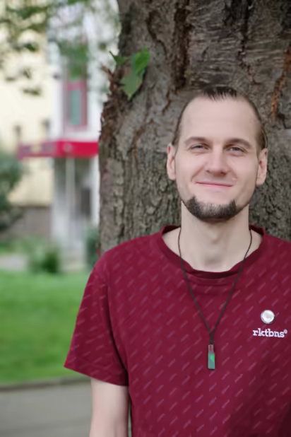 ich, lächelnd, mit zusammengebundenen Haaren, an einem Baum lehnend